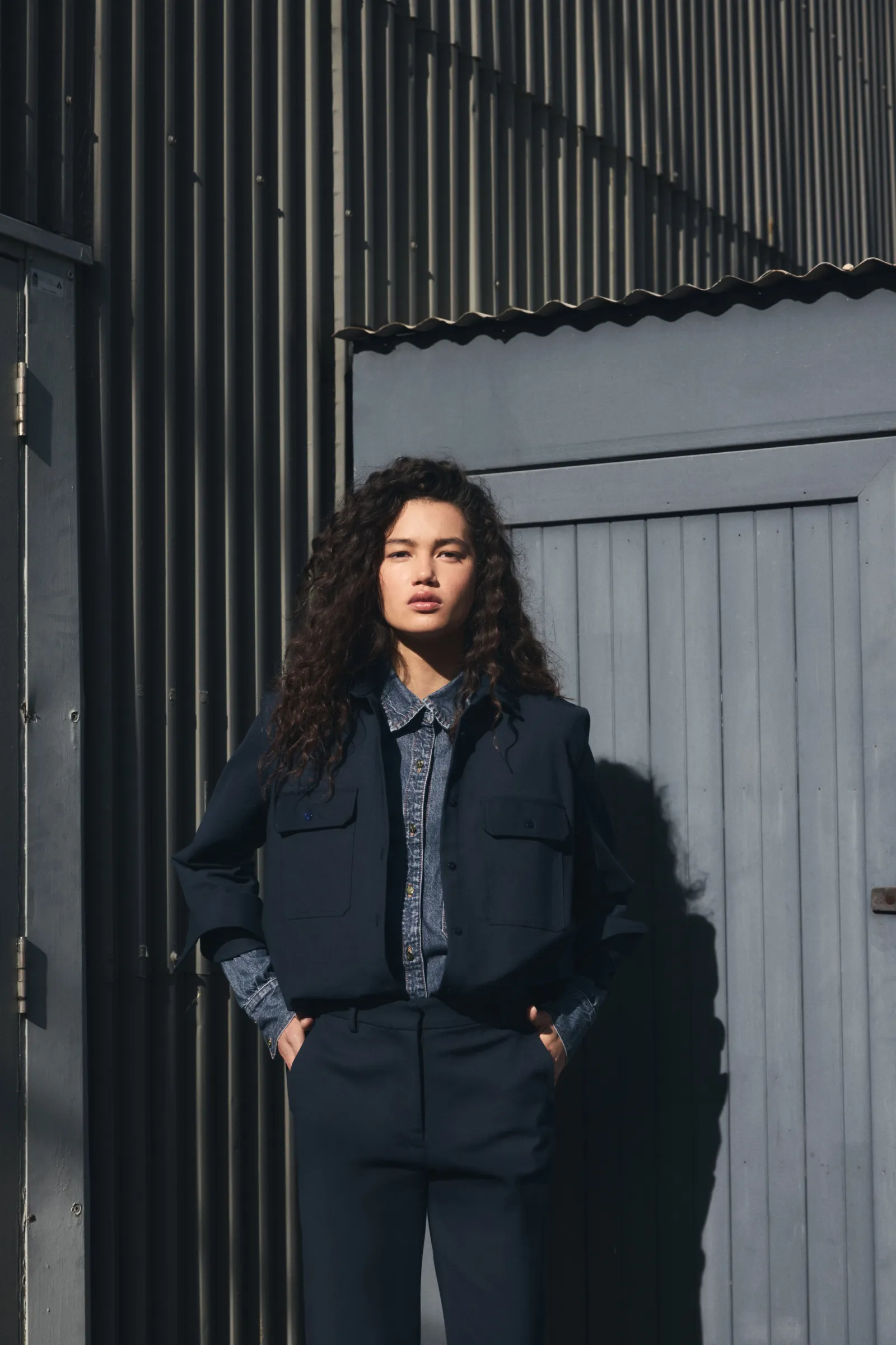 Woman in a dark blue jacket and pants standing outdoors in front of a textured metal wall, posing in soft, natural light.
