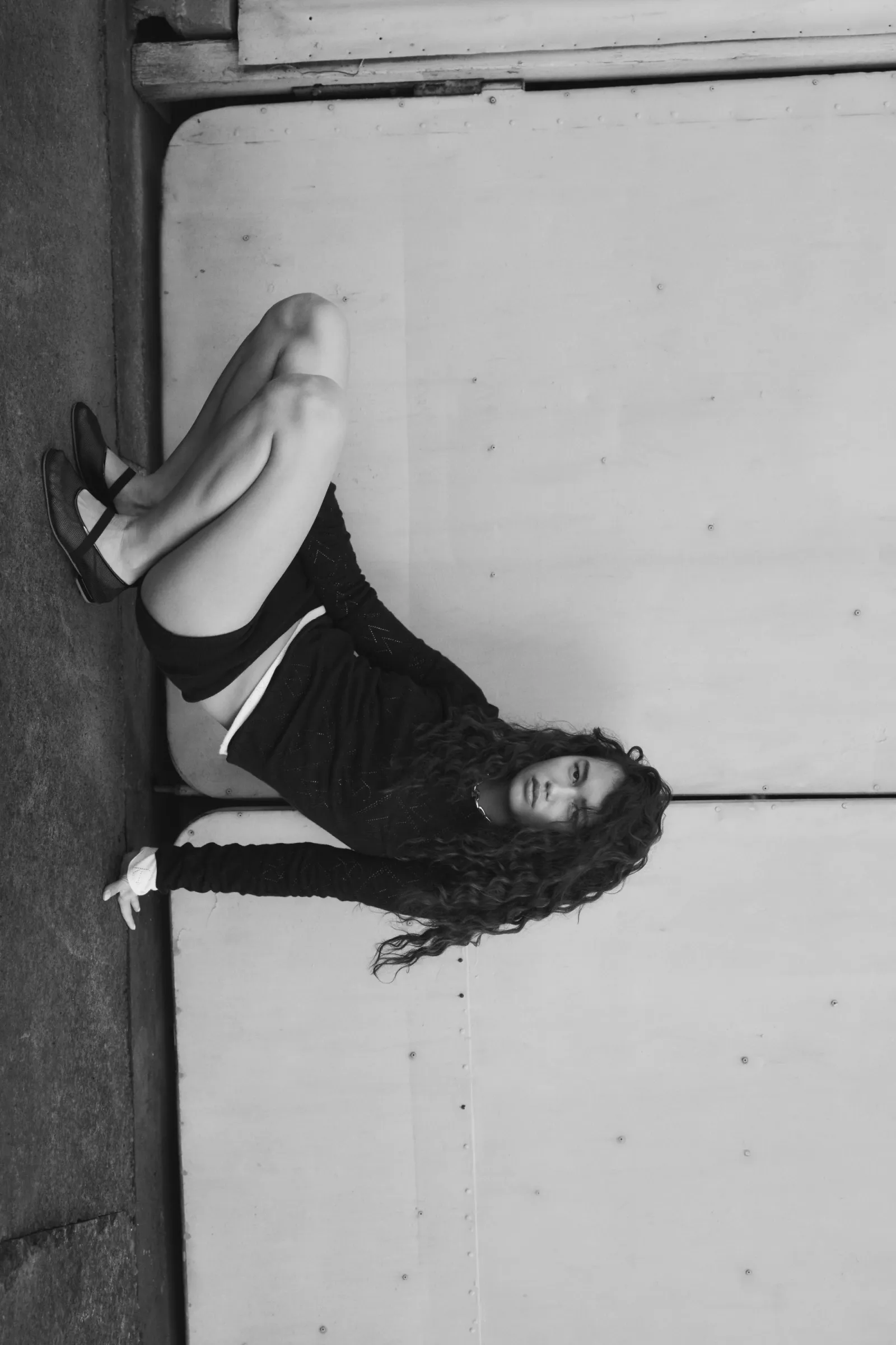 Monochrome portrait of a woman with curly hair in a crouched pose, wearing a black outfit, against a gray textured wall. Fashion photography.