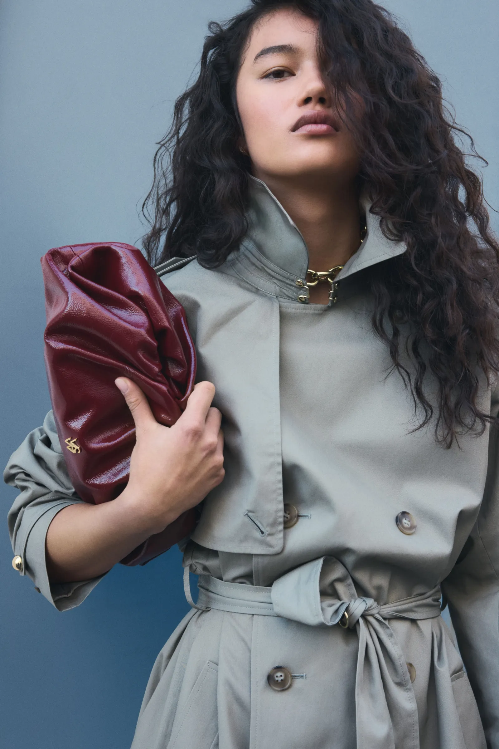 Adult female model poses in gray trench coat holding shiny red clutch purse against a blue background in studio lighting.