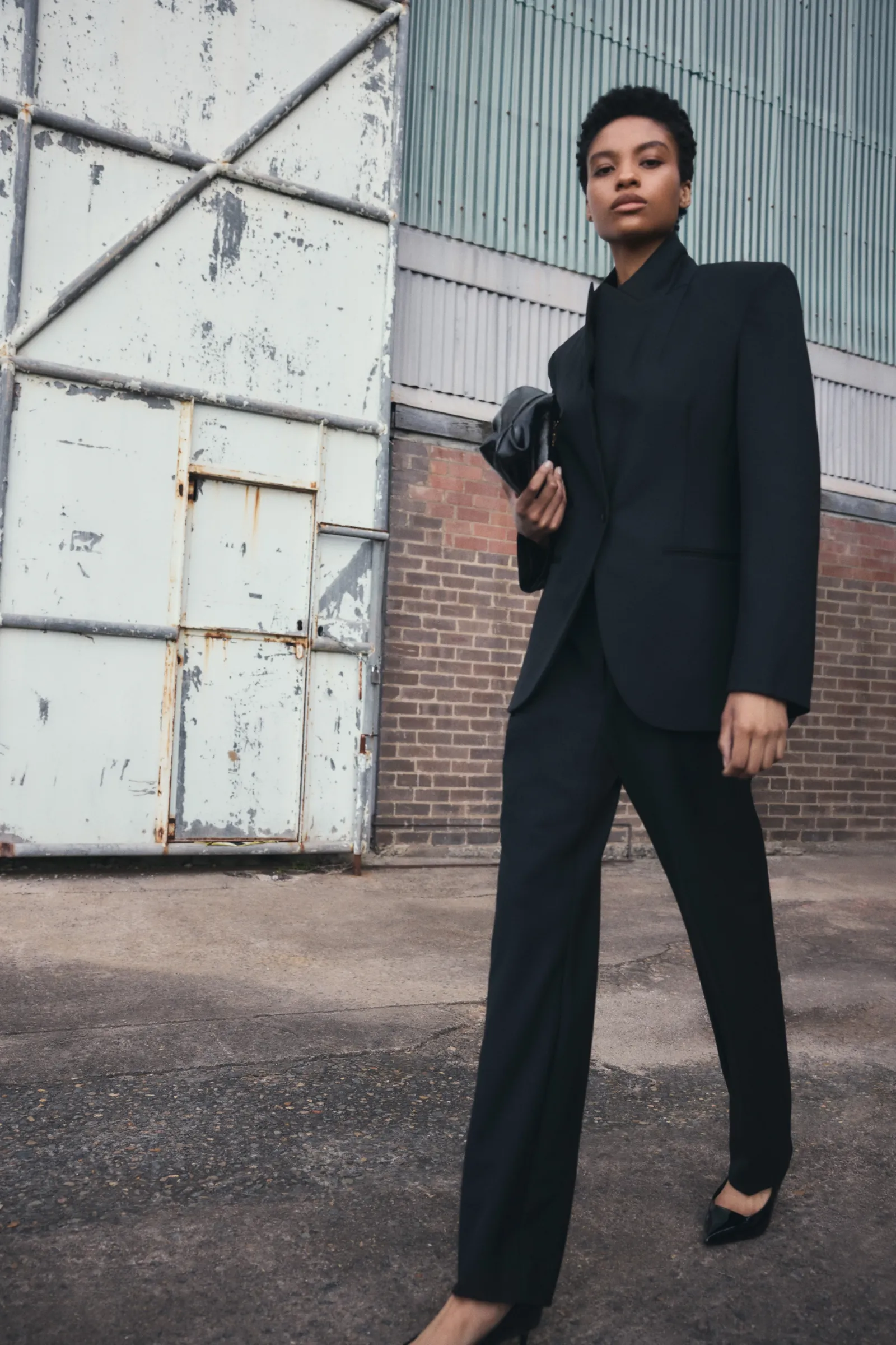 A Black woman in a black blazer and pants poses confidently in front of a warehouse door and brick wall, holding a clutch.