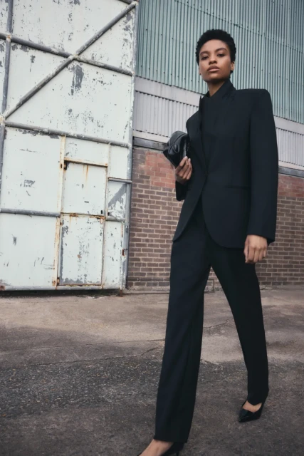 A Black woman in a black blazer and pants poses confidently in front of a warehouse door and brick wall, holding a clutch.
