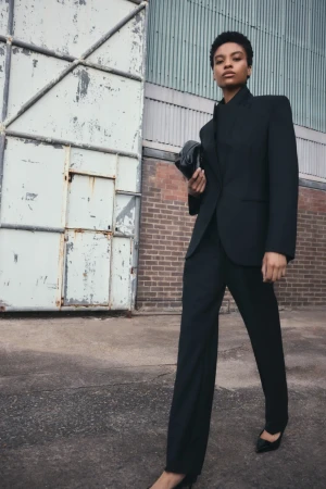 A Black woman in a black blazer and pants poses confidently in front of a warehouse door and brick wall, holding a clutch.