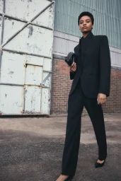 Model in black suit holding a clutch bag walking past a brick wall and industrial door for Aura project