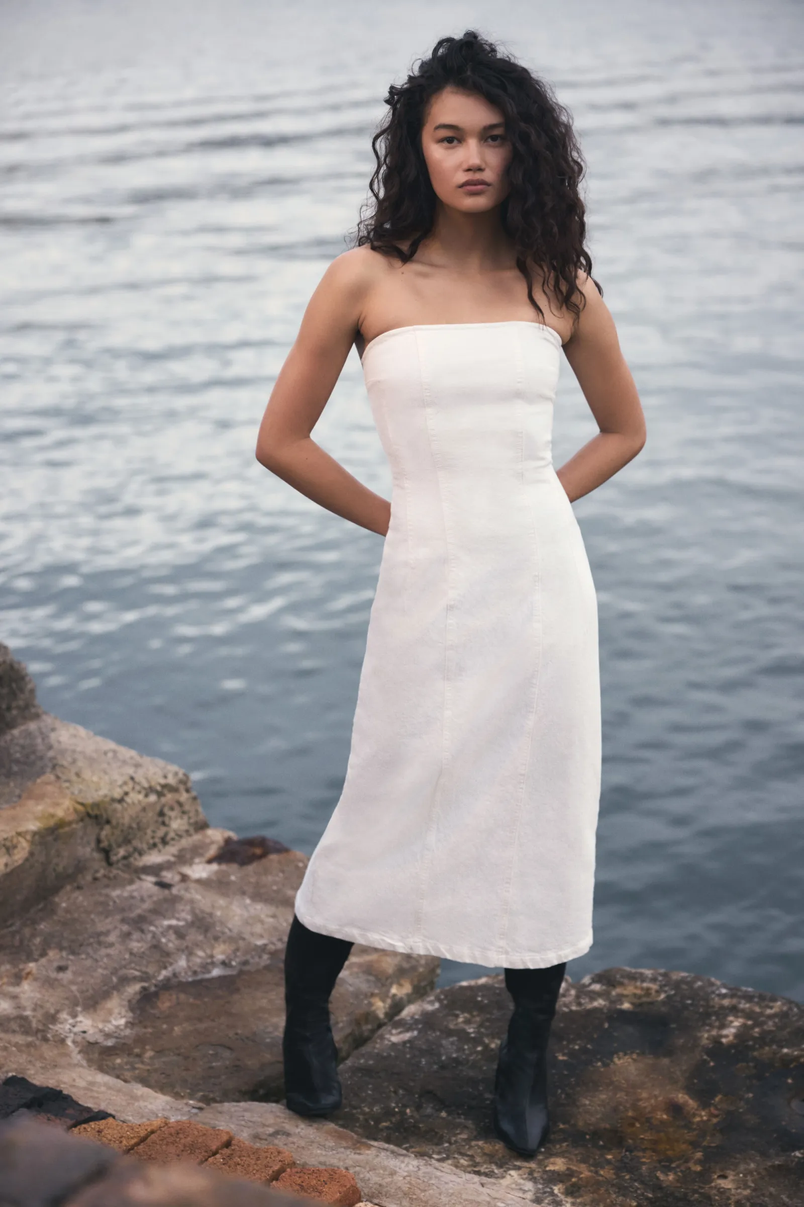 A young woman stands on a rocky surface by the ocean, modeling a white dress and black boots in a fashion shot, capturing a serene mood.