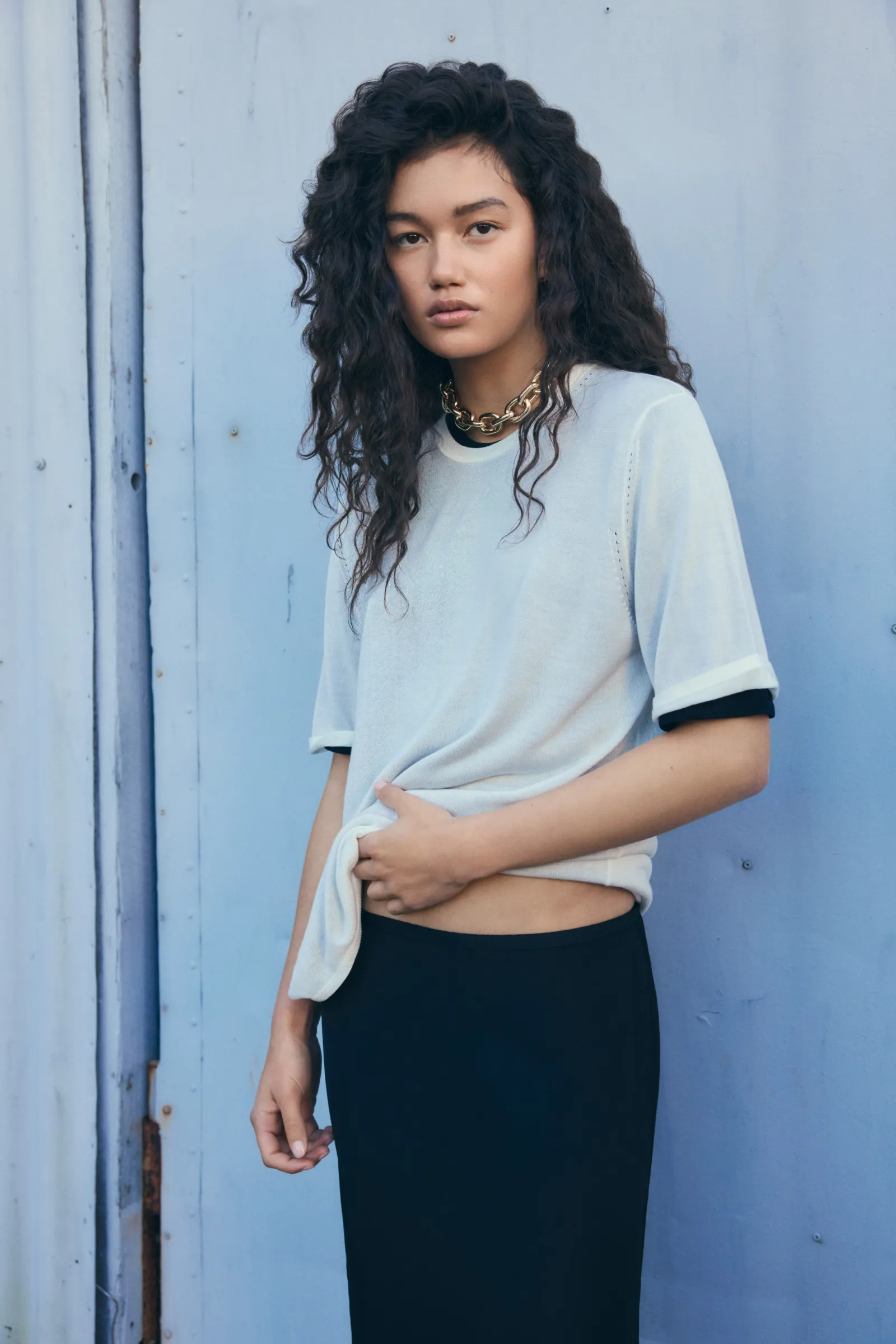 Portrait of a young woman with curly dark hair wearing a T-shirt and skirt, posing against a blue wall in soft, natural light.