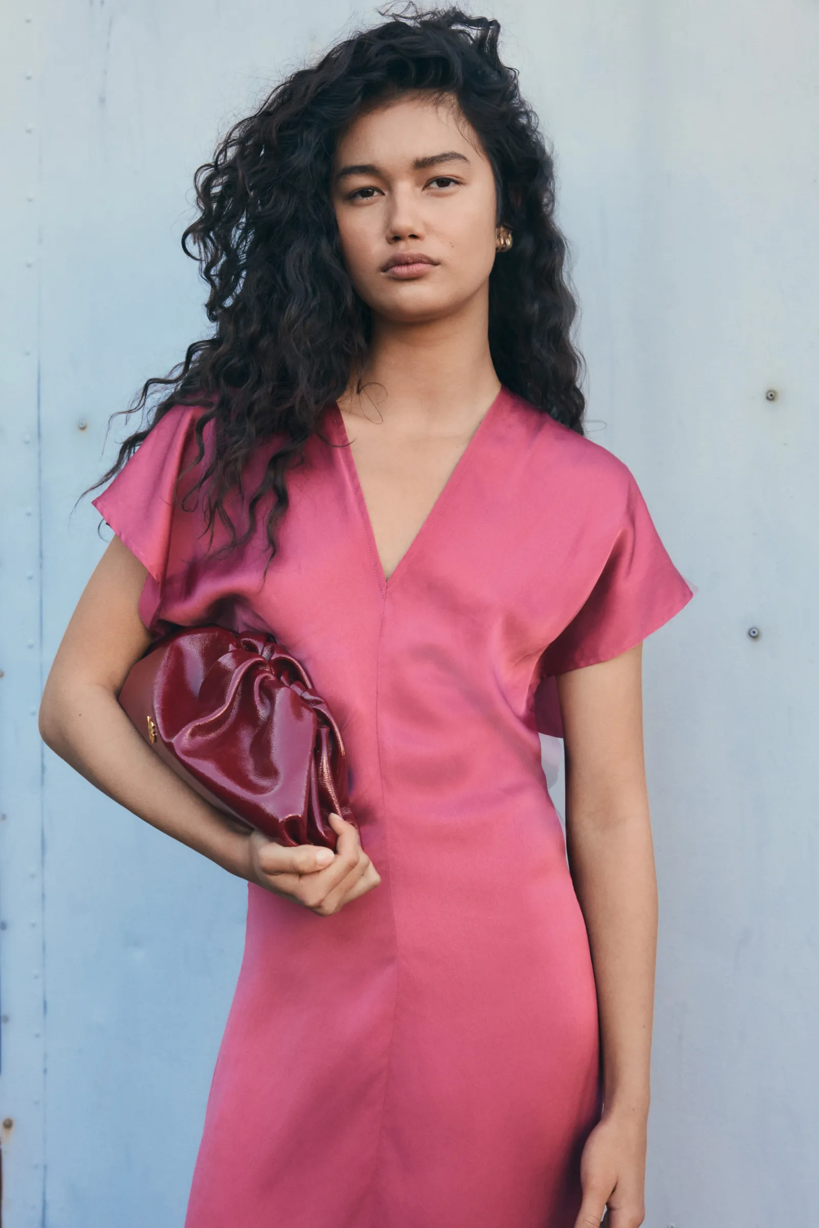A woman poses in a pink V-neck satin dress with short sleeves, holding a glossy maroon clutch, against a muted blue background.