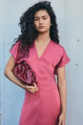 Model with long dark curly hair wearing a pink v-neck dress holding a burgundy clutch bag against a light blue panel wall