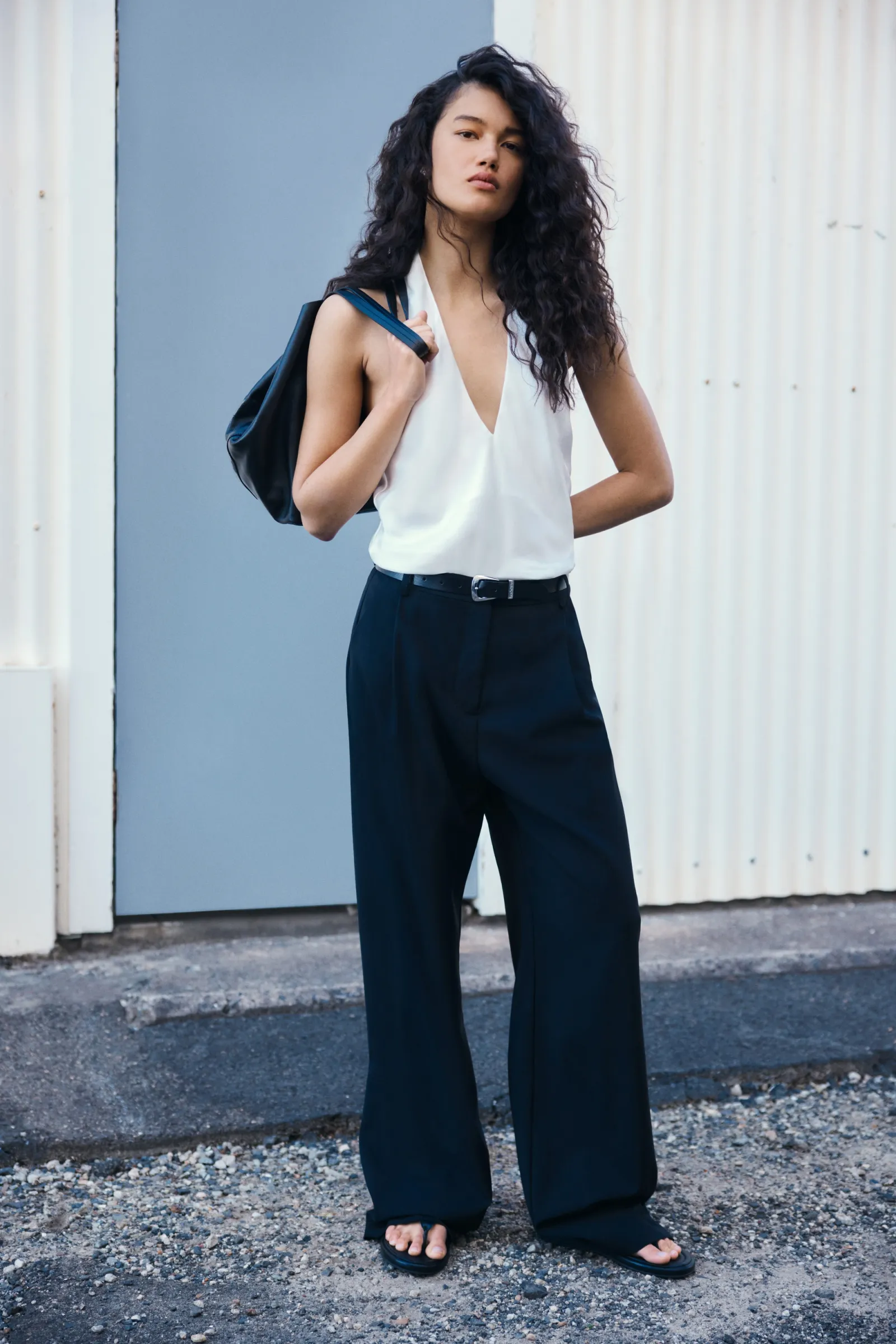 A woman with curly hair models a white top, black pants, and a shoulder bag, standing against a textured wall outdoors in soft daylight.