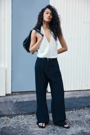 A woman with curly hair models a white top, black pants, and a shoulder bag, standing against a textured wall outdoors in soft daylight.
