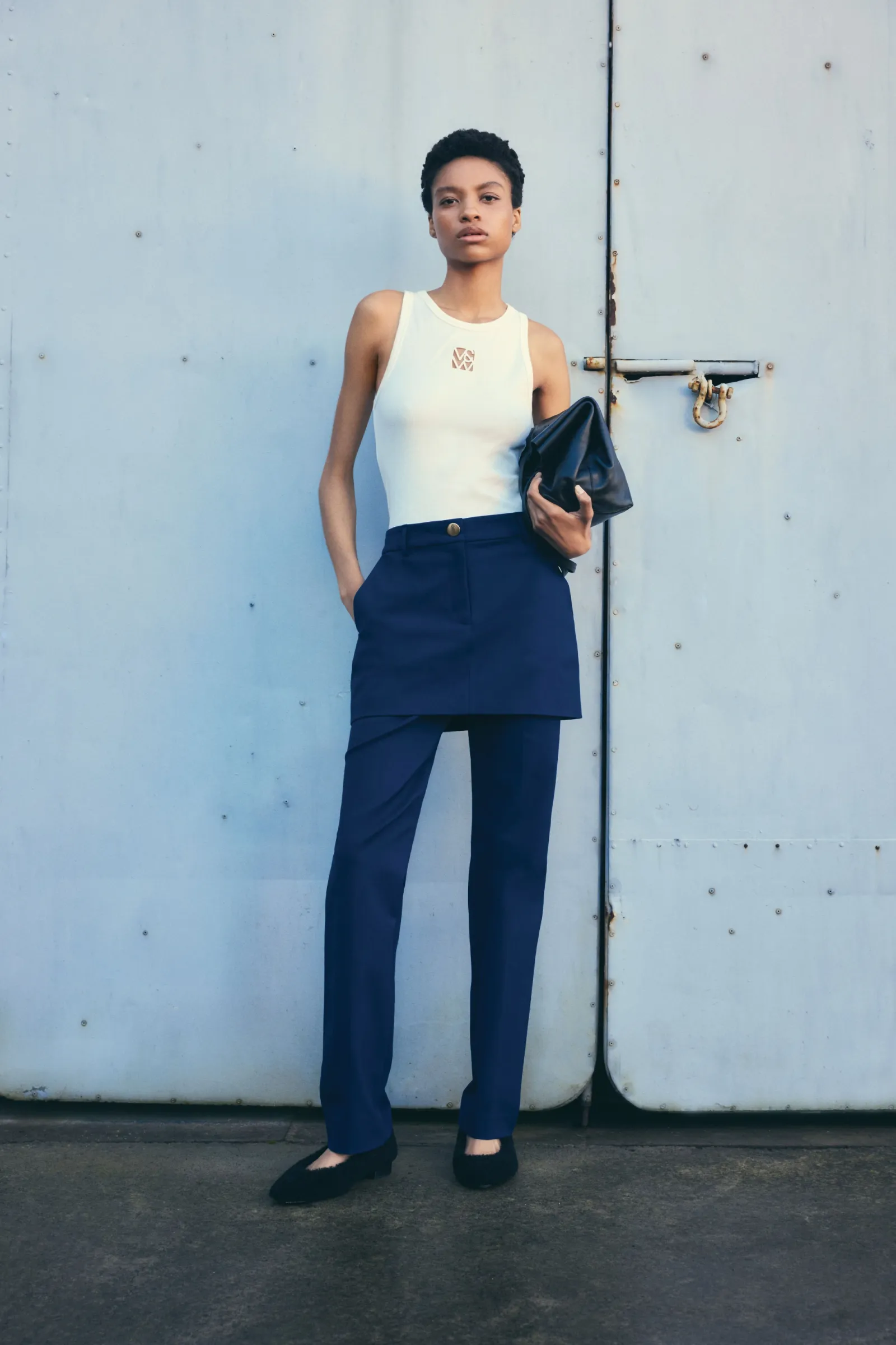 A woman in a cream top, blue pants, and black flats stands in front of a gray metal wall, holding a black bag with both hands.