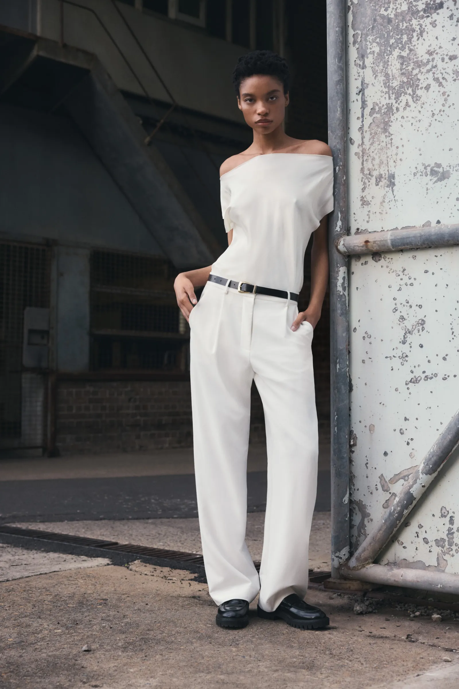 A young adult female poses in a fashion shot wearing a white, off-the-shoulder top and white trousers against a distressed metal background.