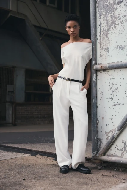 A young adult female poses in a fashion shot wearing a white, off-the-shoulder top and white trousers against a distressed metal background.