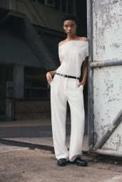 Model in off-the-shoulder white top and white trousers leaning against a large industrial doorway