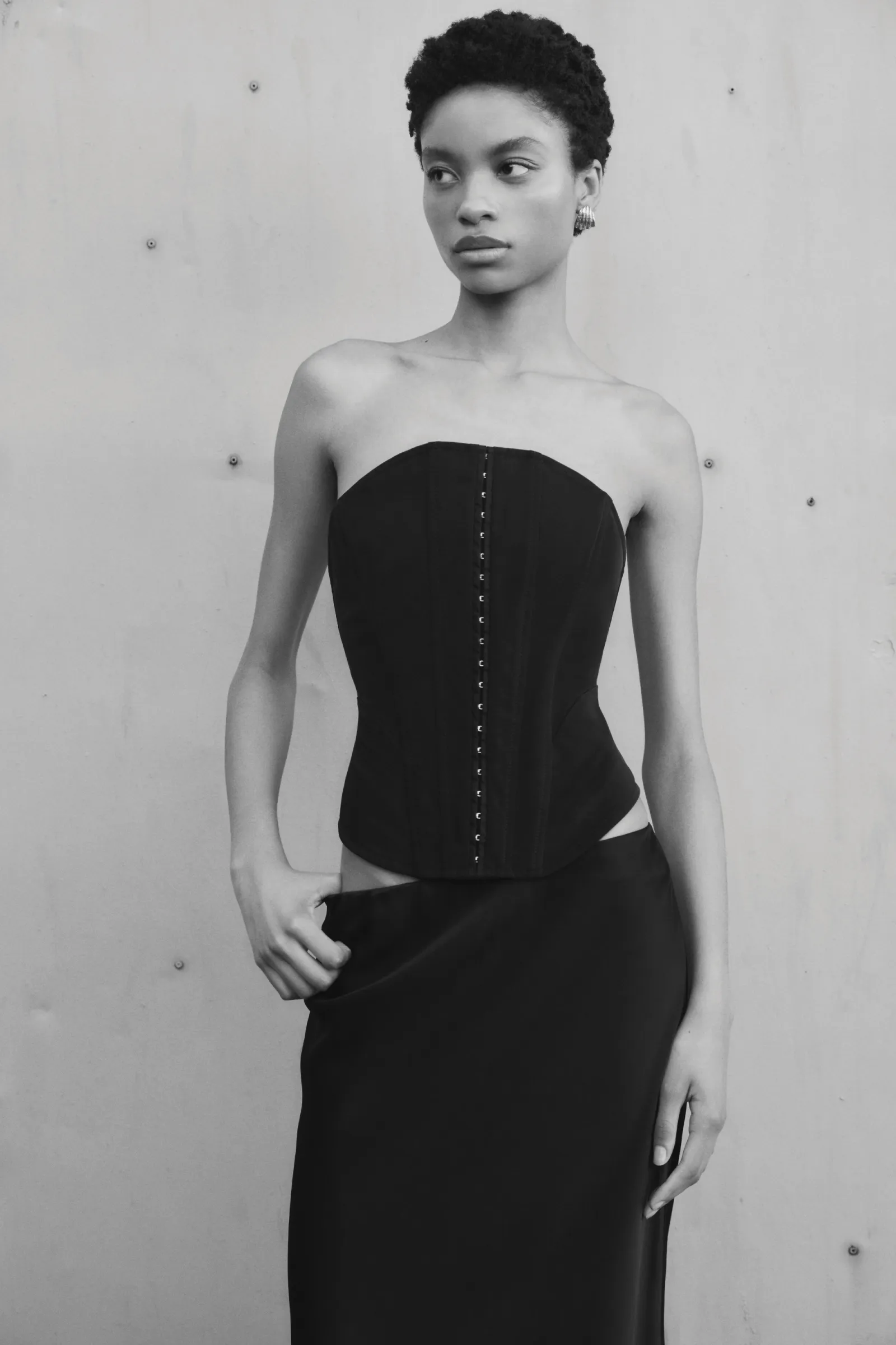 Monochrome portrait of a Black female model in a black corset and skirt against a grey studio backdrop, demonstrating fashion and style.