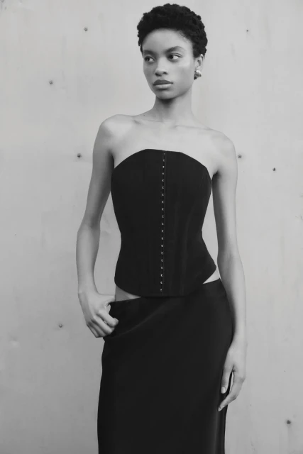 Monochrome portrait of a Black female model in a black corset and skirt against a grey studio backdrop, demonstrating fashion and style.