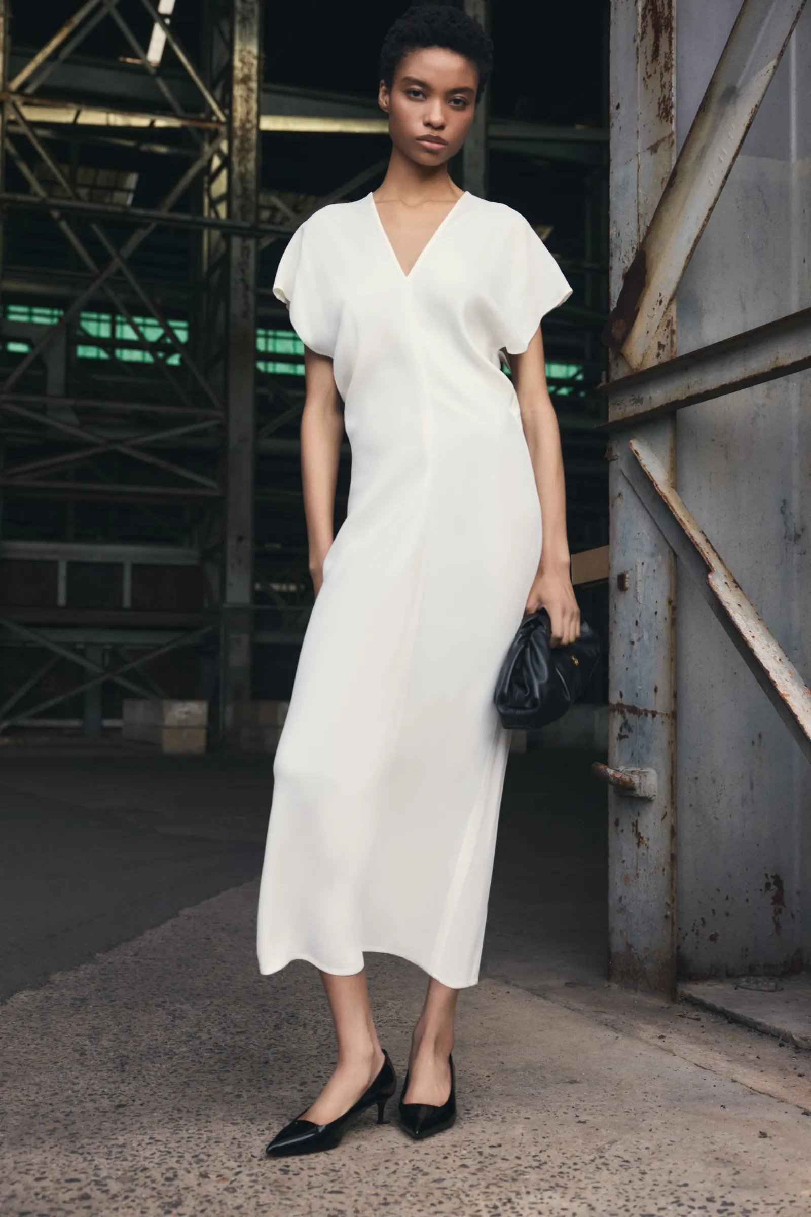 Elegant woman in a cream-colored midi dress poses in front of an industrial backdrop, holding a small black clutch and wearing heeled shoes.