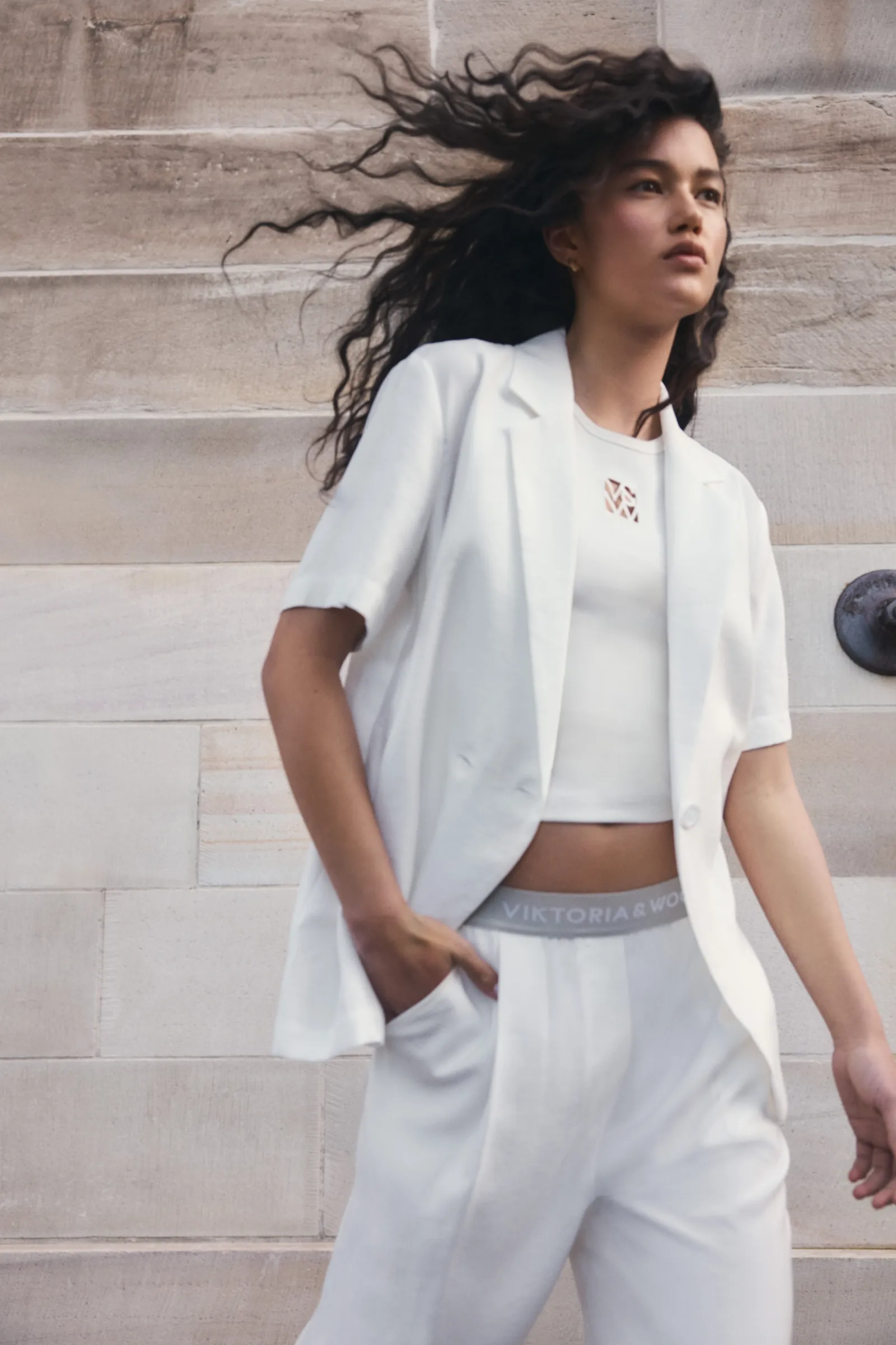 A fashionable young woman models a matching white short-sleeve blazer and pants outfit against a neutral architectural backdrop.