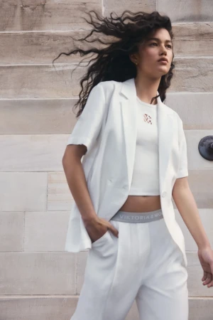 A fashionable young woman models a matching white short-sleeve blazer and pants outfit against a neutral architectural backdrop.