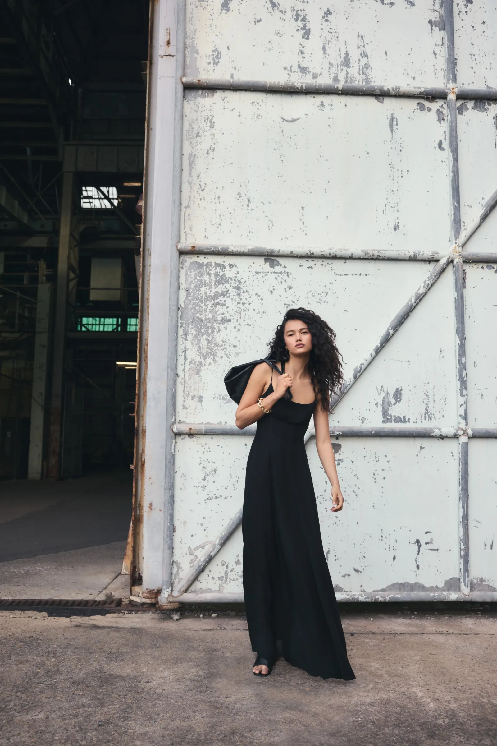 A brunette woman in a long black dress and shoulder bag poses in front of an industrial warehouse door. Fashion editorial, daytime shot.