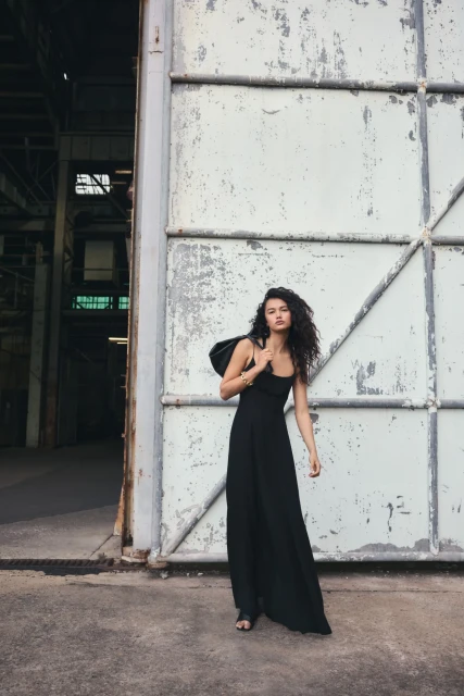 A brunette woman in a long black dress and shoulder bag poses in front of an industrial warehouse door. Fashion editorial, daytime shot.