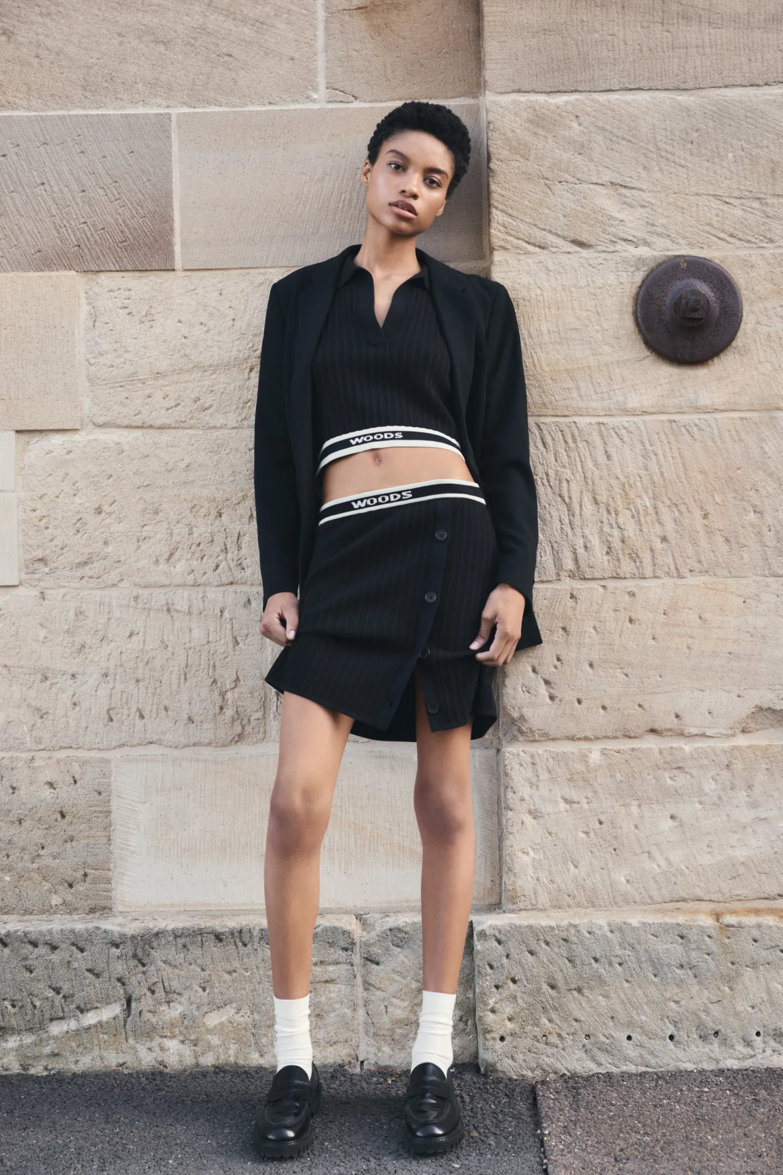 An adult female poses against a textured stone wall, wearing a black top, skirt, blazer, socks, and shoes in a medium shot.