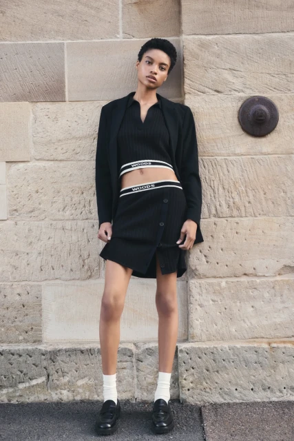 An adult female poses against a textured stone wall, wearing a black top, skirt, blazer, socks, and shoes in a medium shot.
