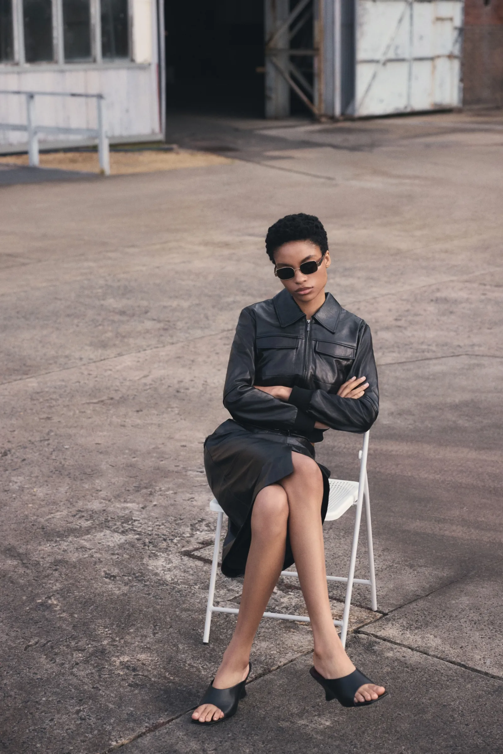 Woman in black leather jacket and skirt sits on white folding chair outdoors. Wearing sunglasses and black heeled sandals.