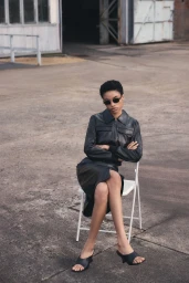Model seated on a white folding chair outdoors wearing a black leather jacket and matching skirt with sunglasses