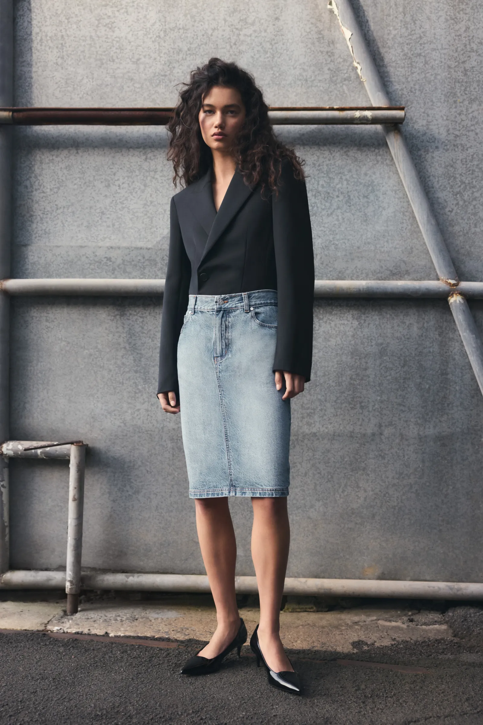 A woman poses in a black blazer and denim skirt against a gray industrial backdrop. She is wearing black heels and has curly hair.