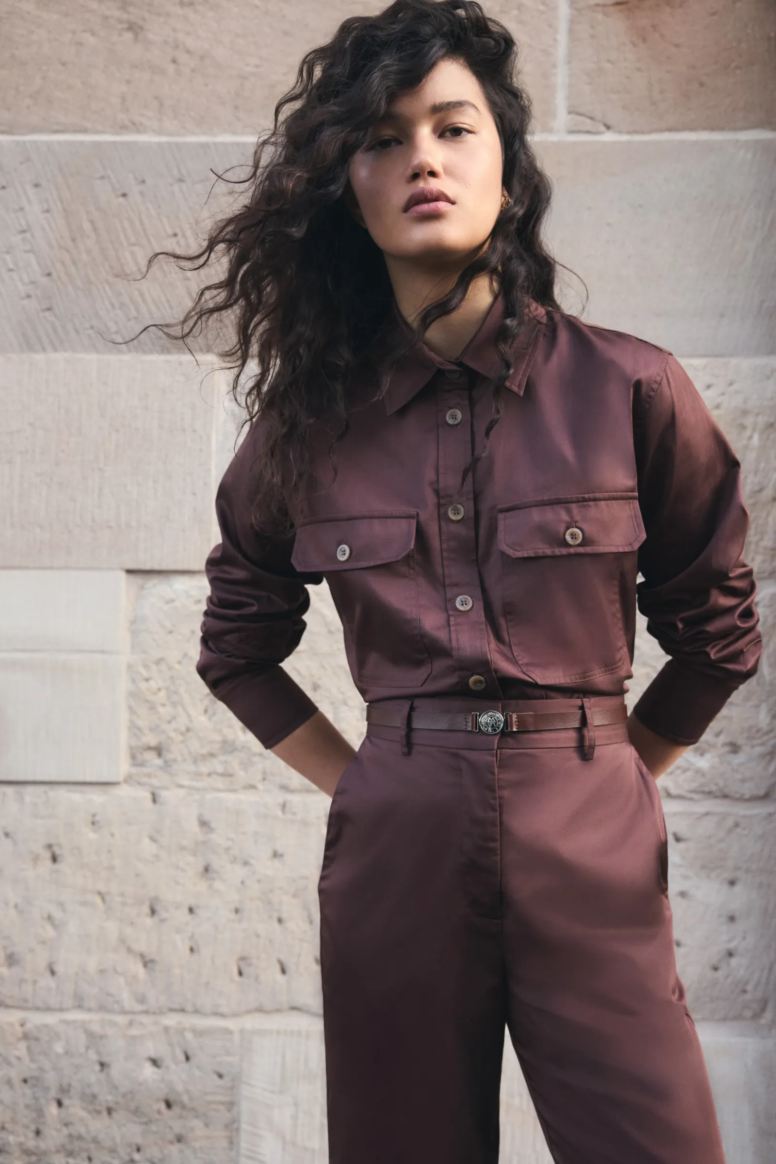 A woman in a brown two-piece shirt and pants stands in front of a stone wall, looking directly at the camera in a medium shot.