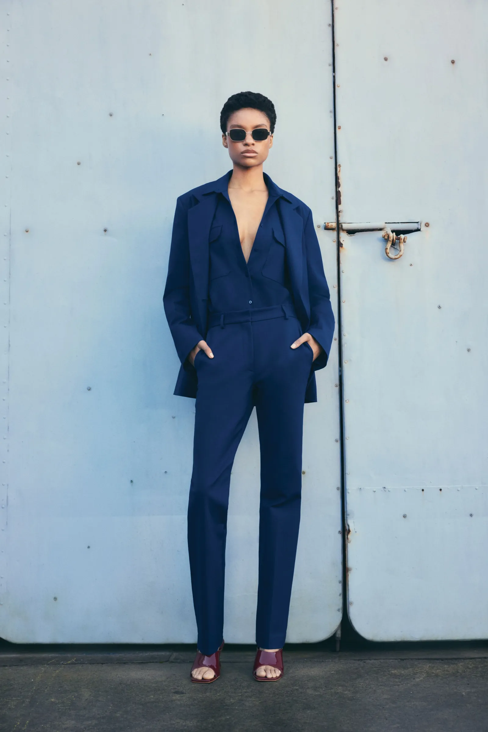 Fashion shot of a woman in a dark blue pantsuit and sunglasses standing against a weathered metal wall, showcasing modern elegance.