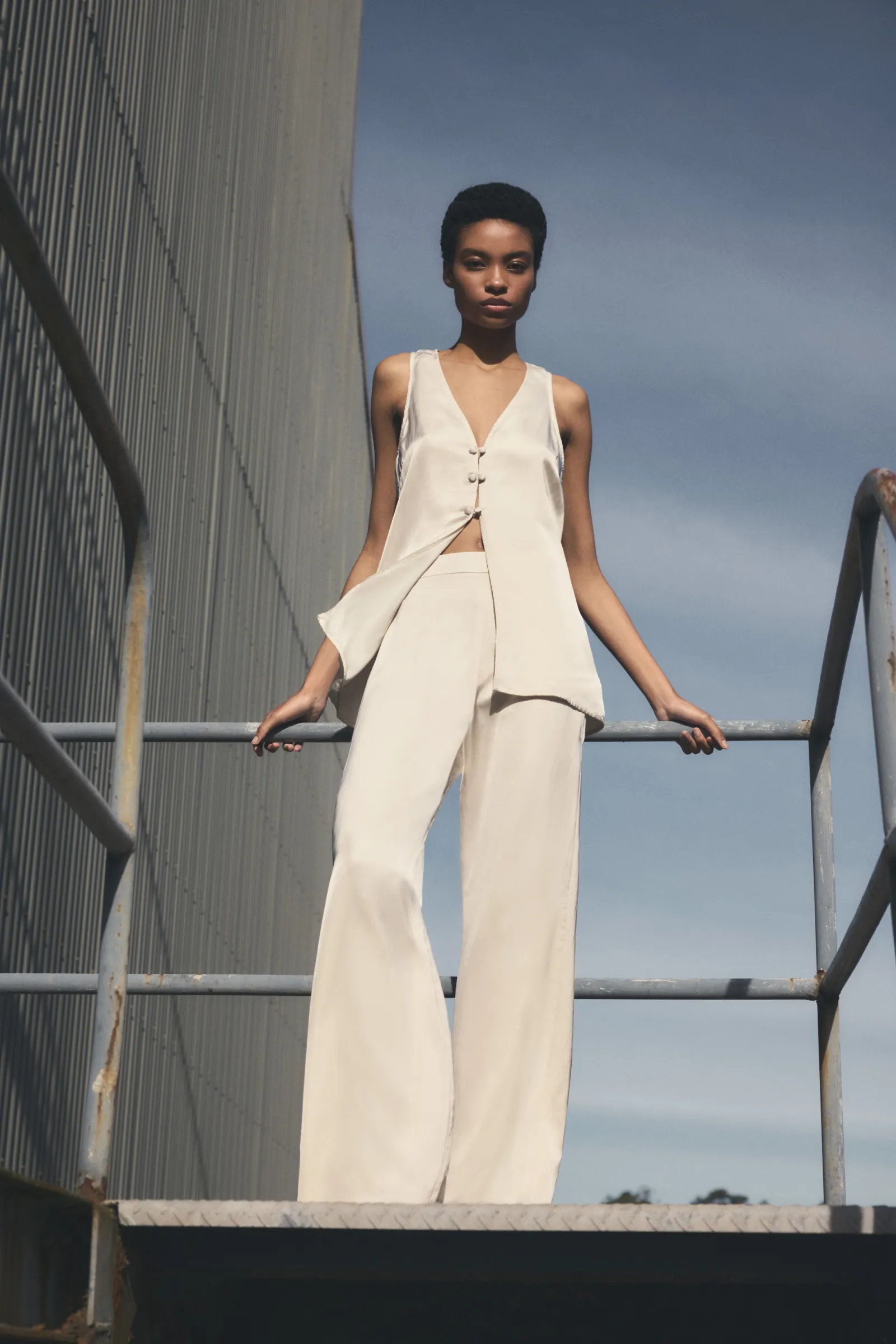 A woman in a sleeveless top and pants stands on a metal staircase against an industrial building under a clear sky, fashion shoot.