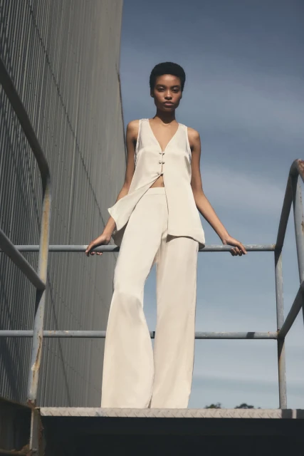 A woman in a sleeveless top and pants stands on a metal staircase against an industrial building under a clear sky, fashion shoot.