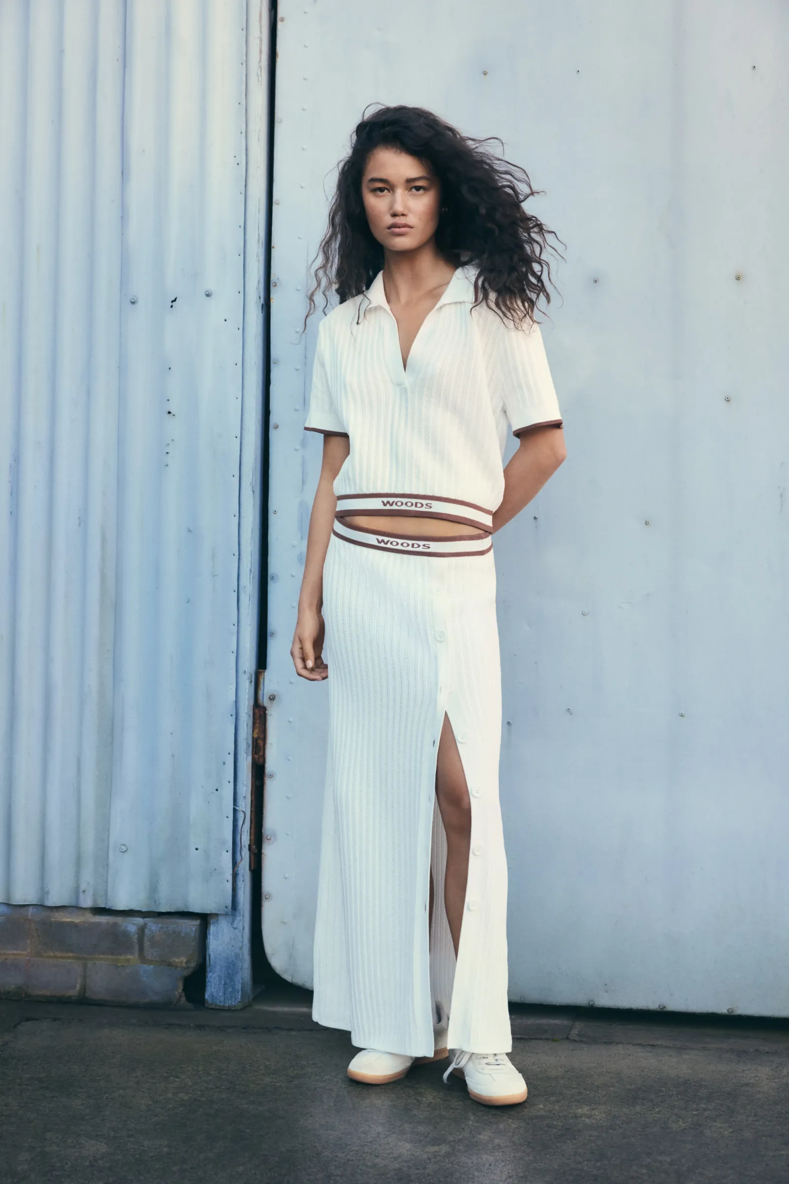 A slender woman with dark curly hair poses in a white, ribbed, high-slit skirt and matching collared top against a blue metal wall.