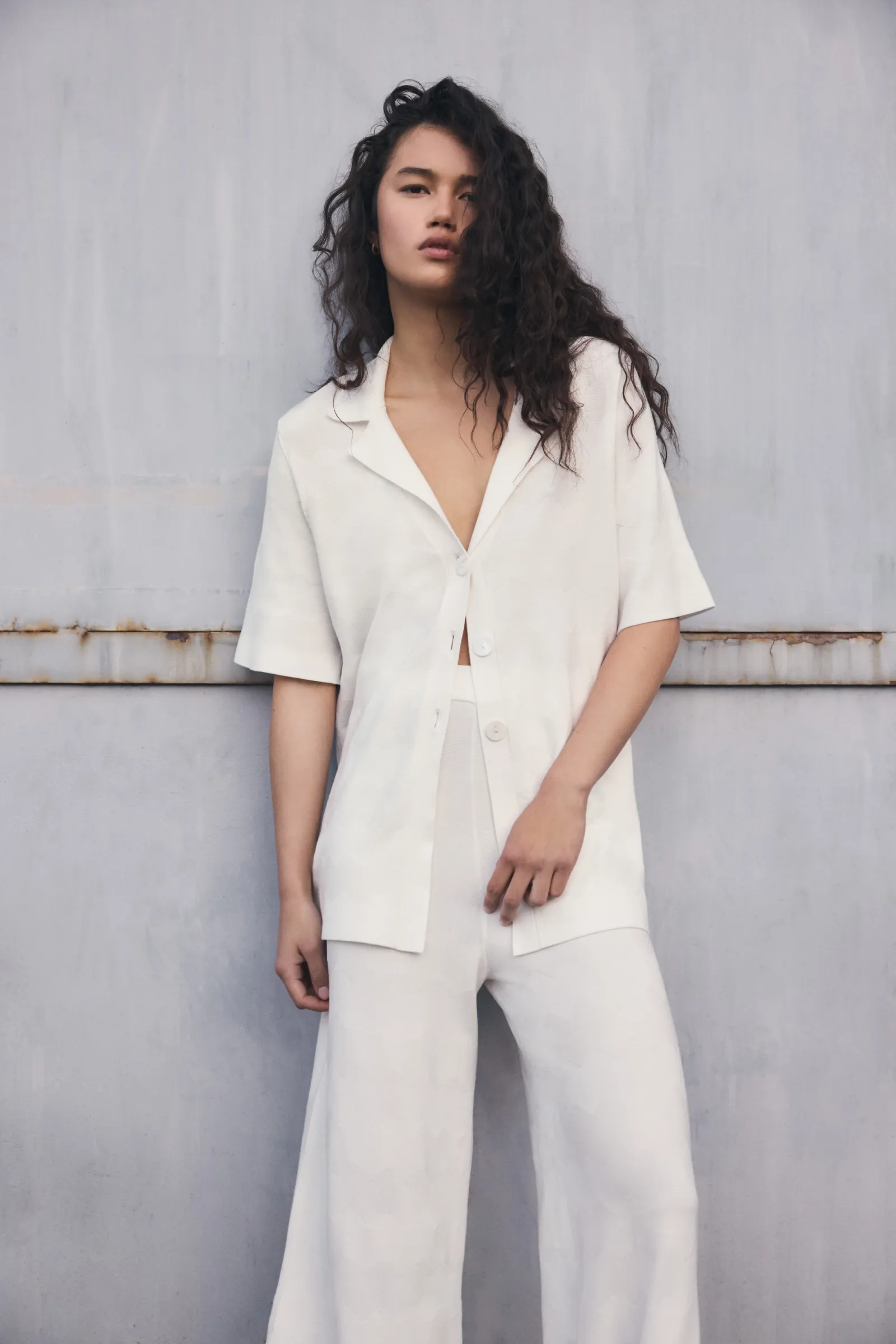 A woman with curly hair poses in an unbuttoned short-sleeve shirt and wide-leg pants, showcasing modern and casual fashion in a studio setting.