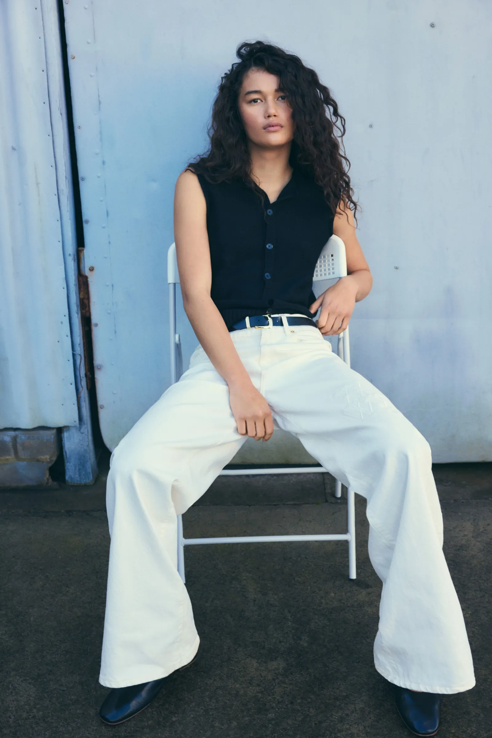Portrait of a woman sitting on a chair wearing a black top and white pants in front of a weathered blue metal wall.