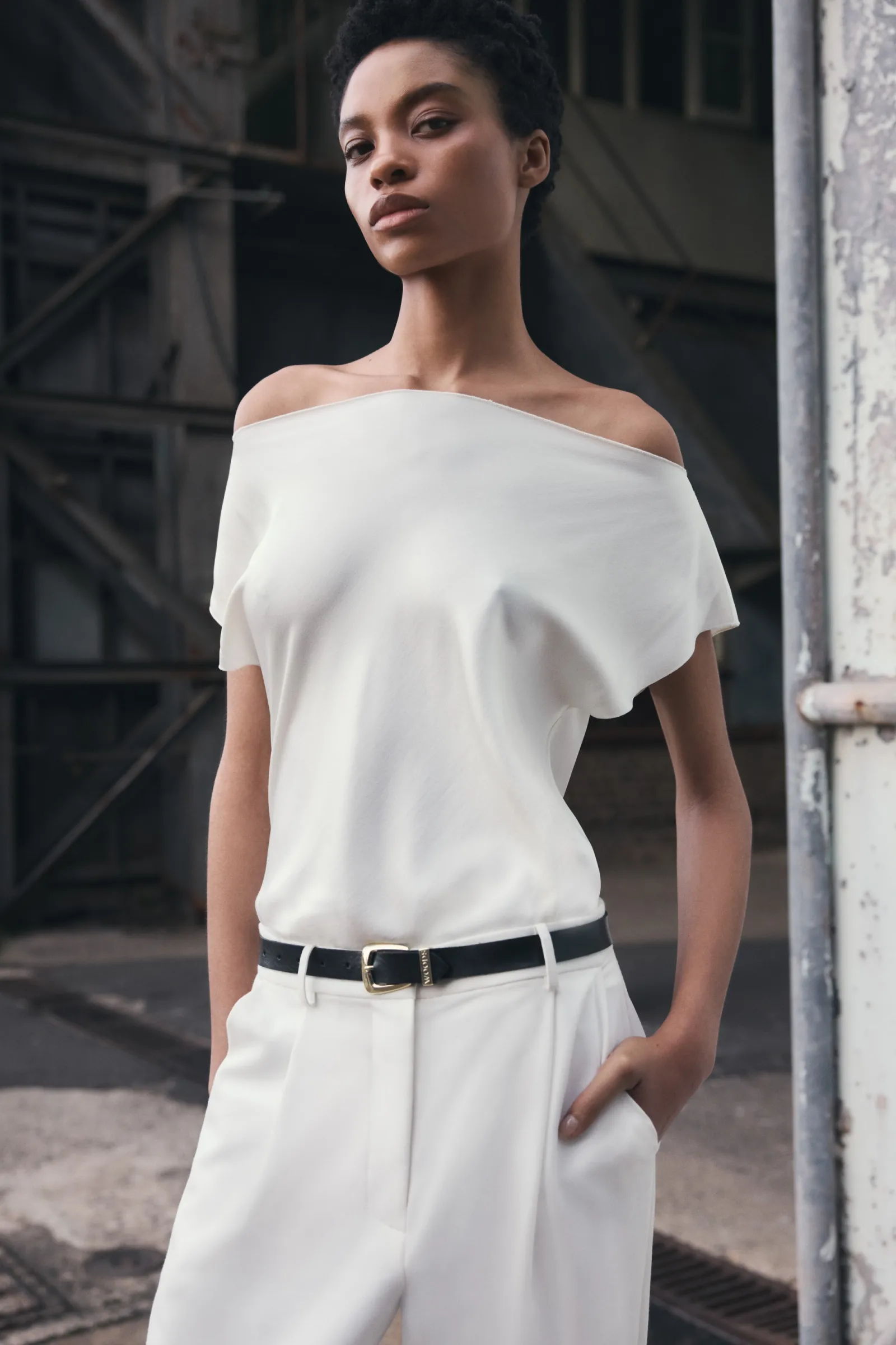 Portrait of a slender woman in a casual white off-the-shoulder top and trousers, standing against an industrial backdrop for a fashion shot.