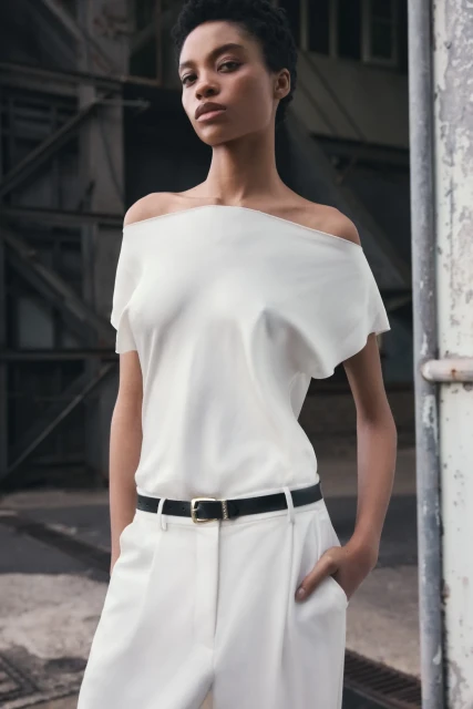 Portrait of a slender woman in a casual white off-the-shoulder top and trousers, standing against an industrial backdrop for a fashion shot.