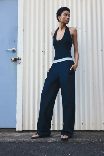 A slender Black woman poses in wide-leg trousers and a halter top against a corrugated metal wall and blue door in an outdoor setting.