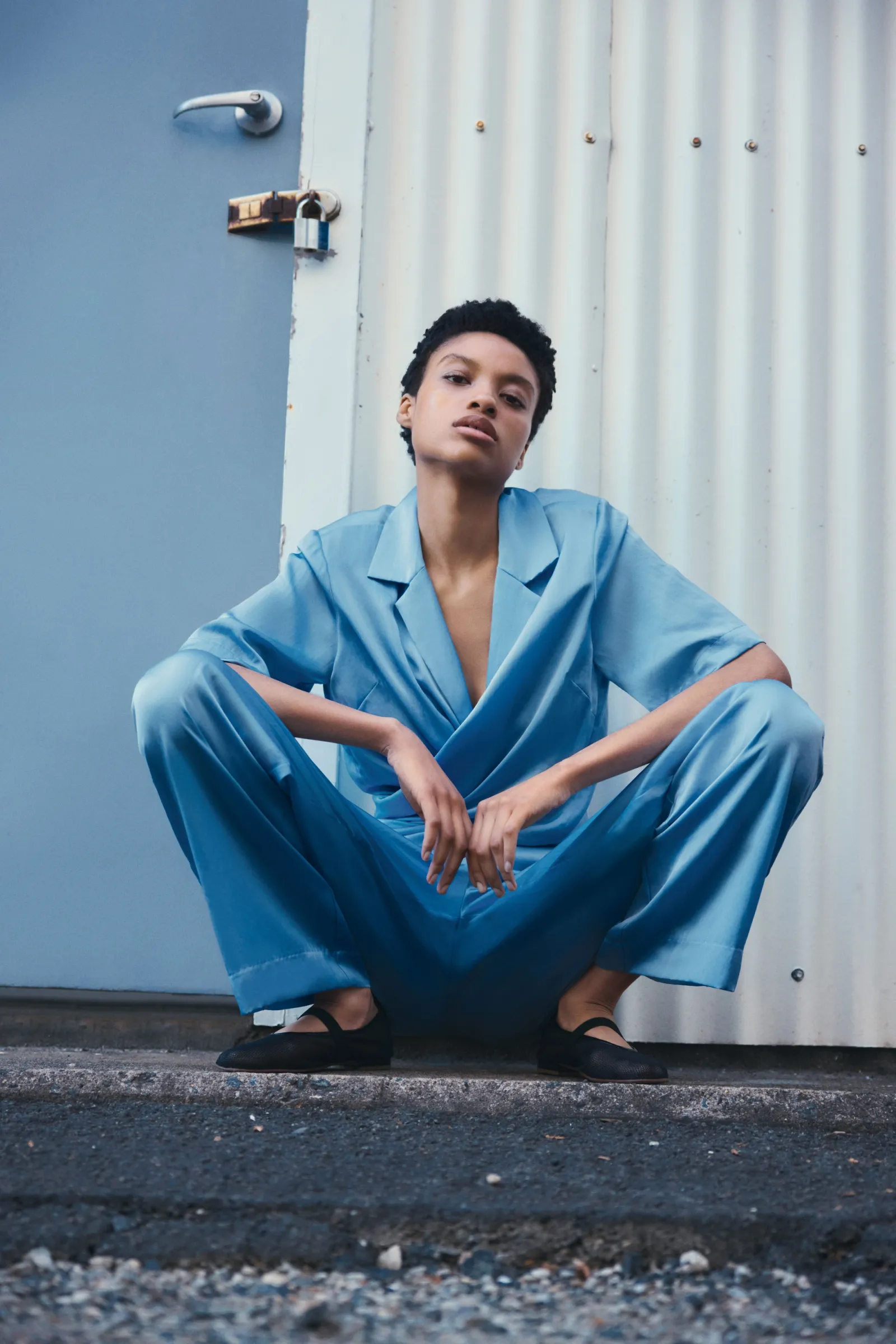 Woman in light blue pantsuit squats outdoors against a white metal wall, looking at the camera. Black shoes and casual attire.