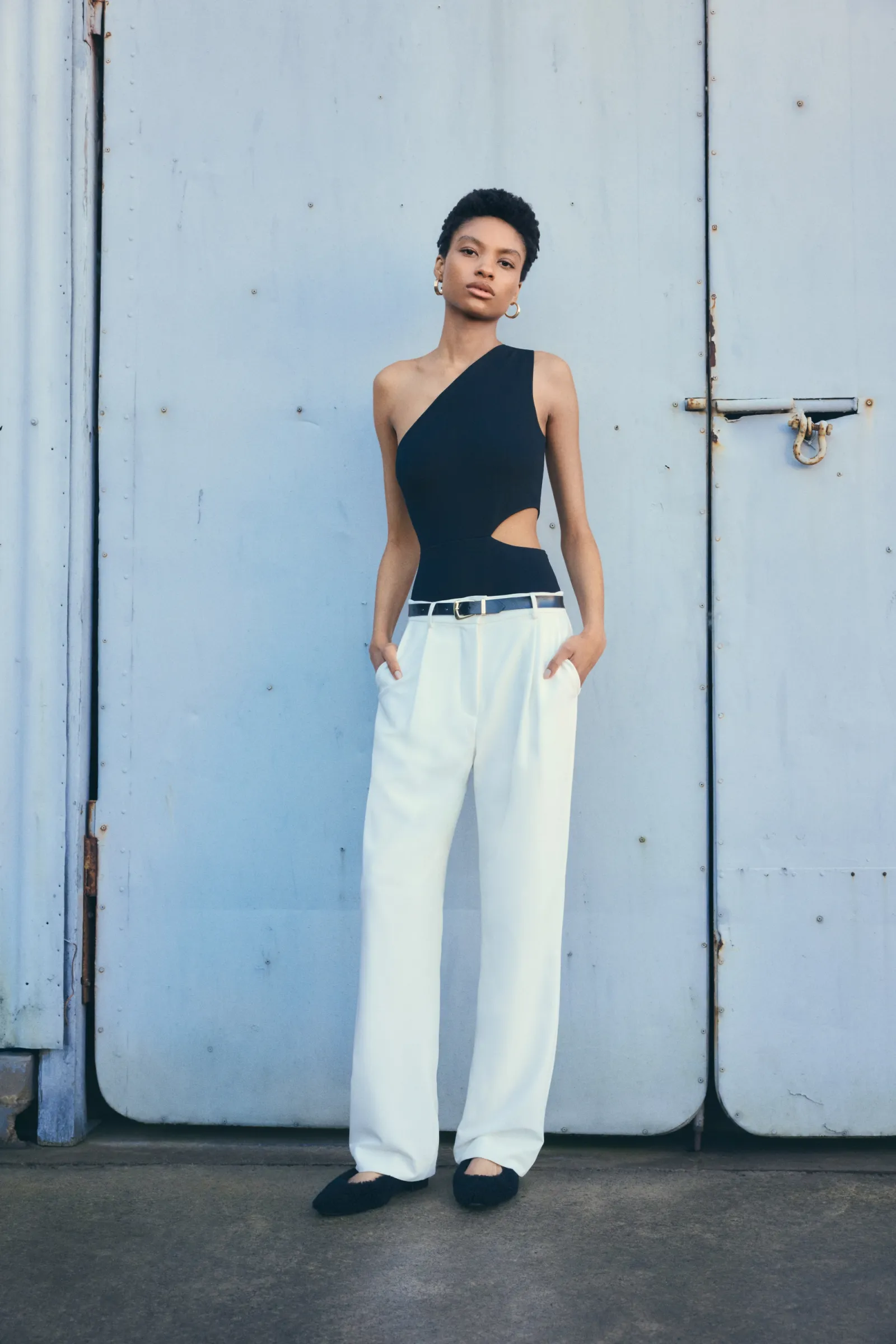 Fashion shot of a Black woman in a black one-shoulder bodysuit and white pants standing against a light blue corrugated wall.