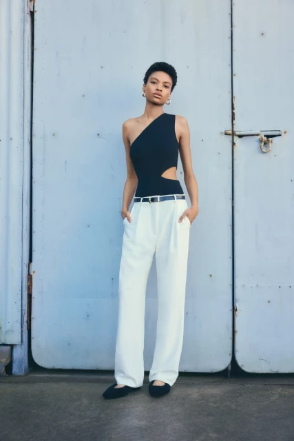 Fashion shot of a Black woman in a black one-shoulder bodysuit and white pants standing against a light blue corrugated wall.