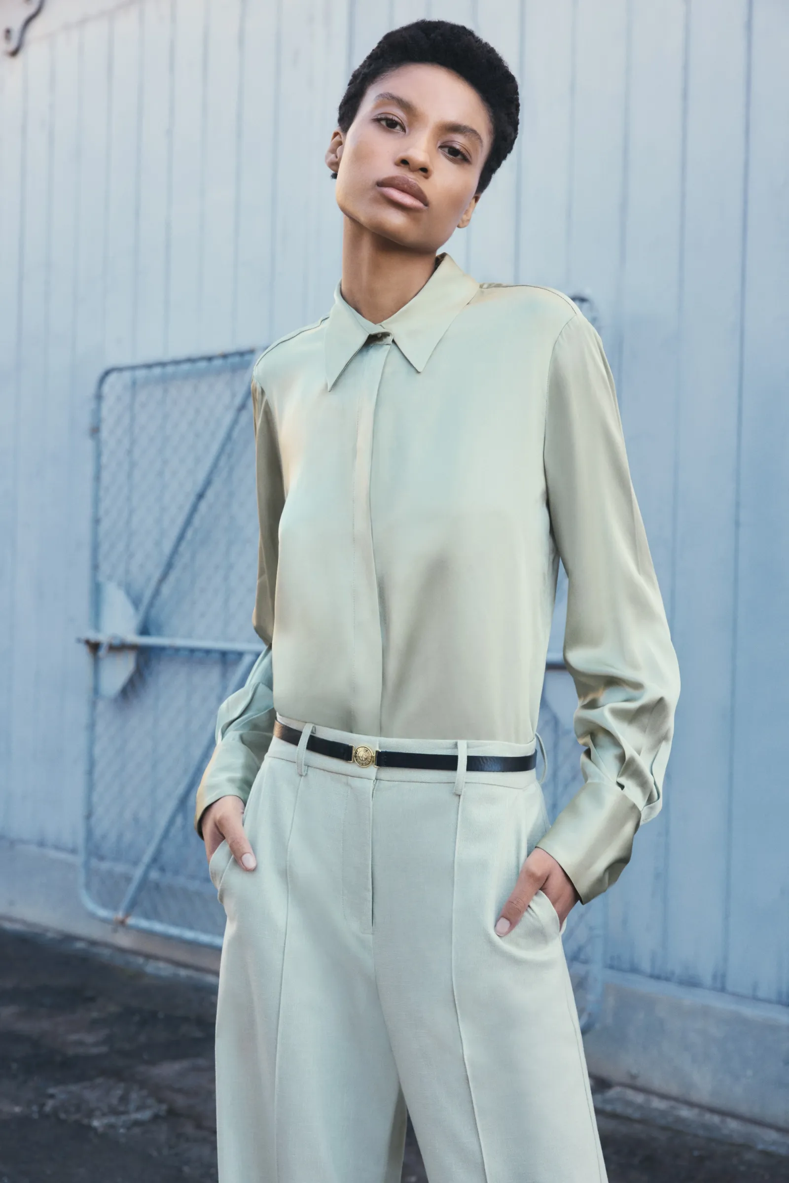 A person in a light green button-down shirt and pants poses in front of a pale blue wall with a metal grate and an urban setting.
