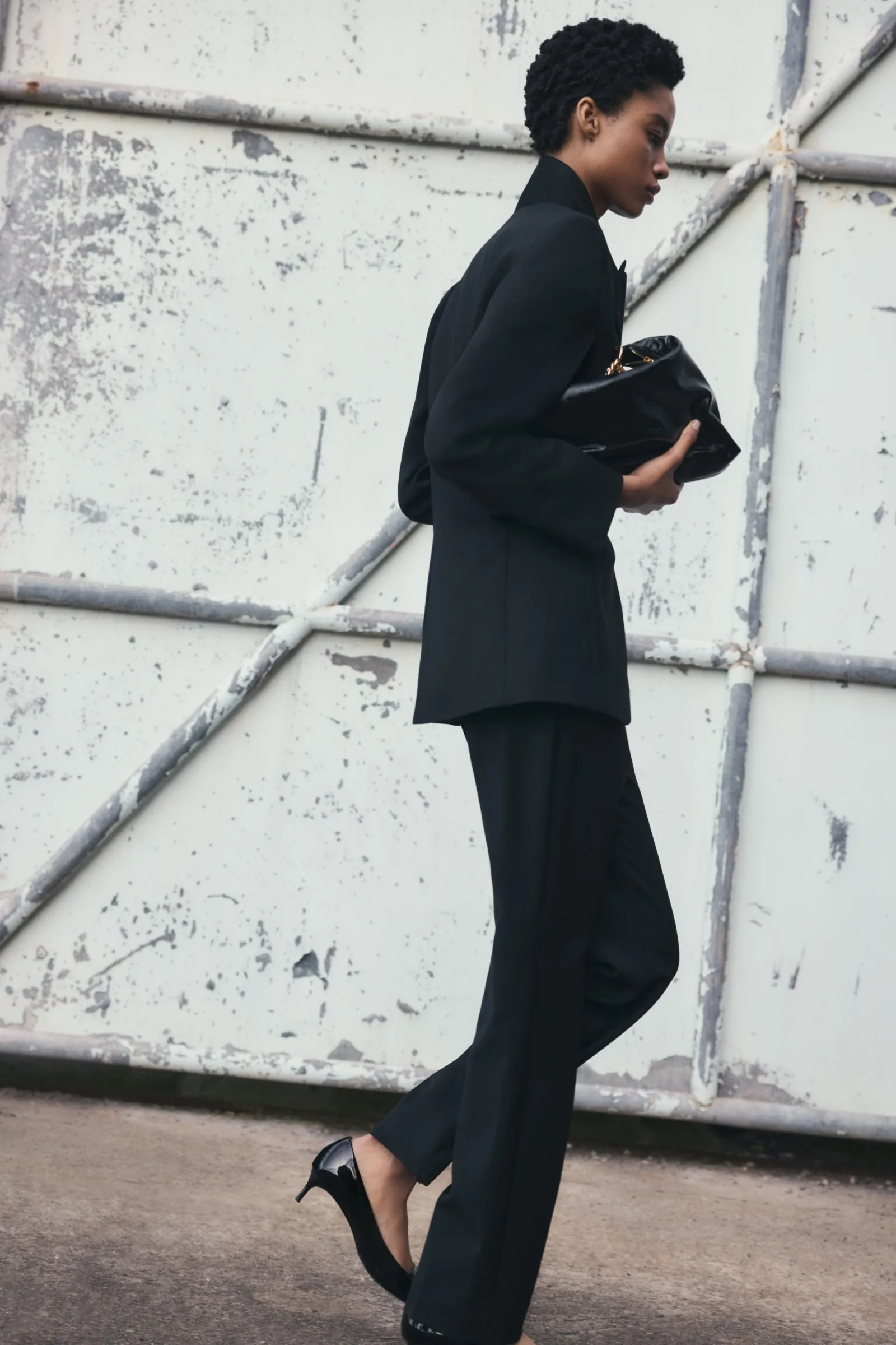 Woman in black suit and heels walks past weathered industrial door, holding a black clutch bag. Fashion editorial, outdoor setting.