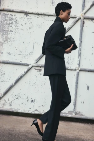 Woman in black suit and heels walks past weathered industrial door, holding a black clutch bag. Fashion editorial, outdoor setting.