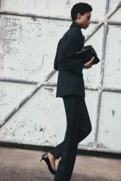 Model in black suit holding a black clutch standing against an industrial wall
