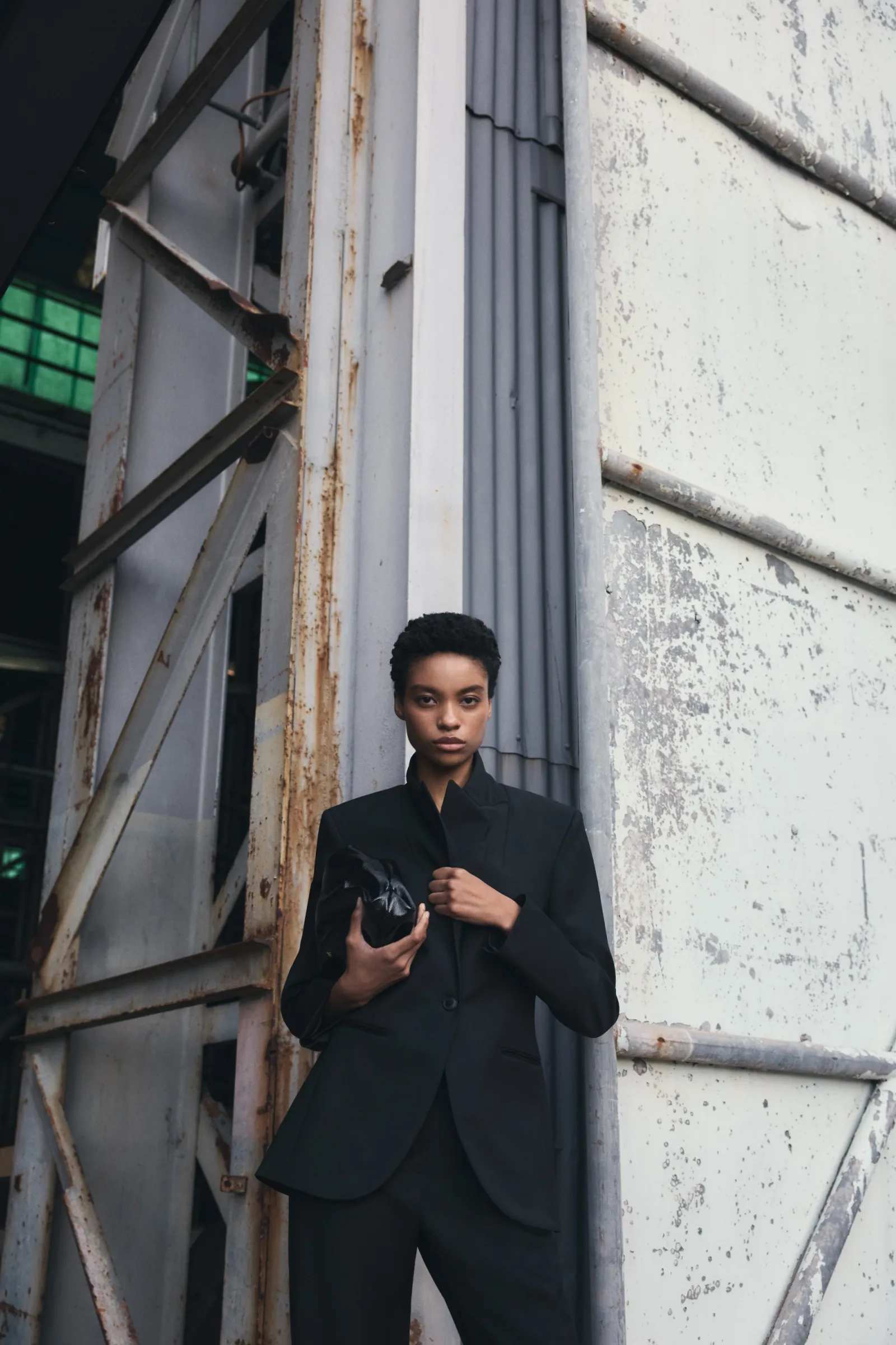 A short-haired woman poses in a black suit with a black clutch, standing against a textured metal and concrete industrial backdrop.
