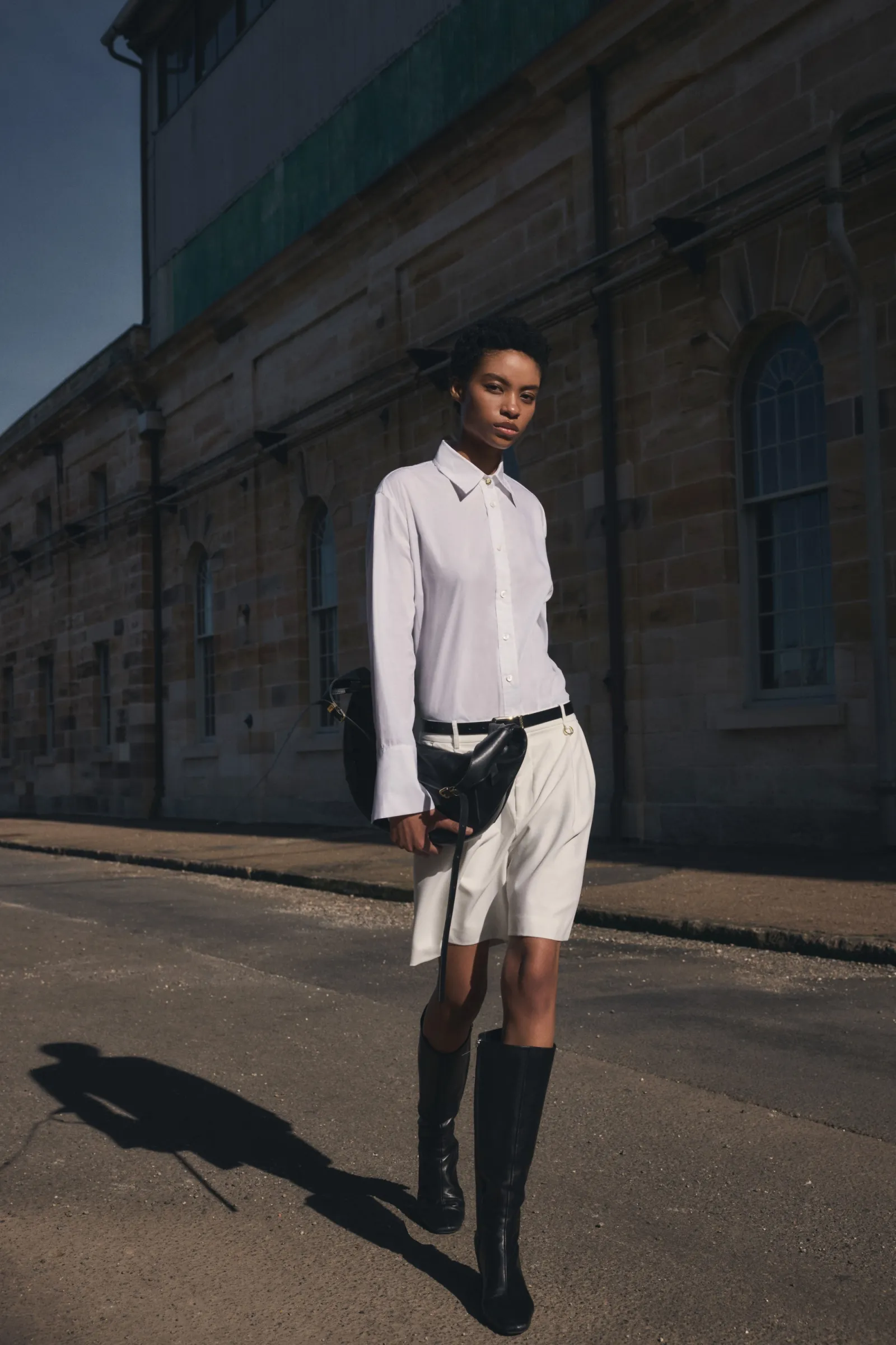 A woman in a white button-down shirt and shorts stands on an urban street in black boots near an industrial building.