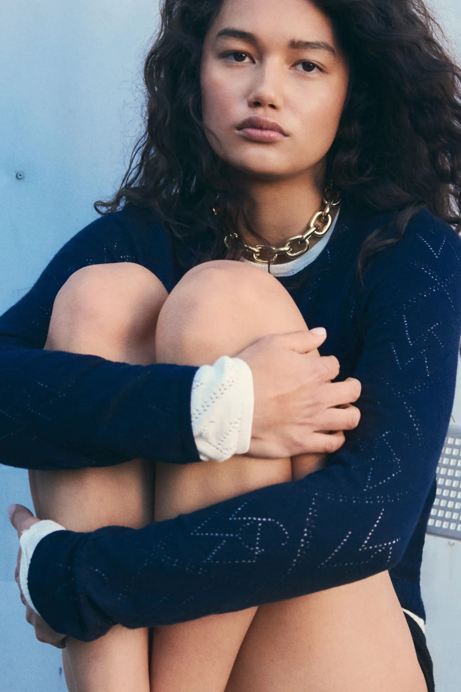A young woman with dark curly hair sits with her arms around her knees, gazing directly at the camera, wearing a navy sweater.