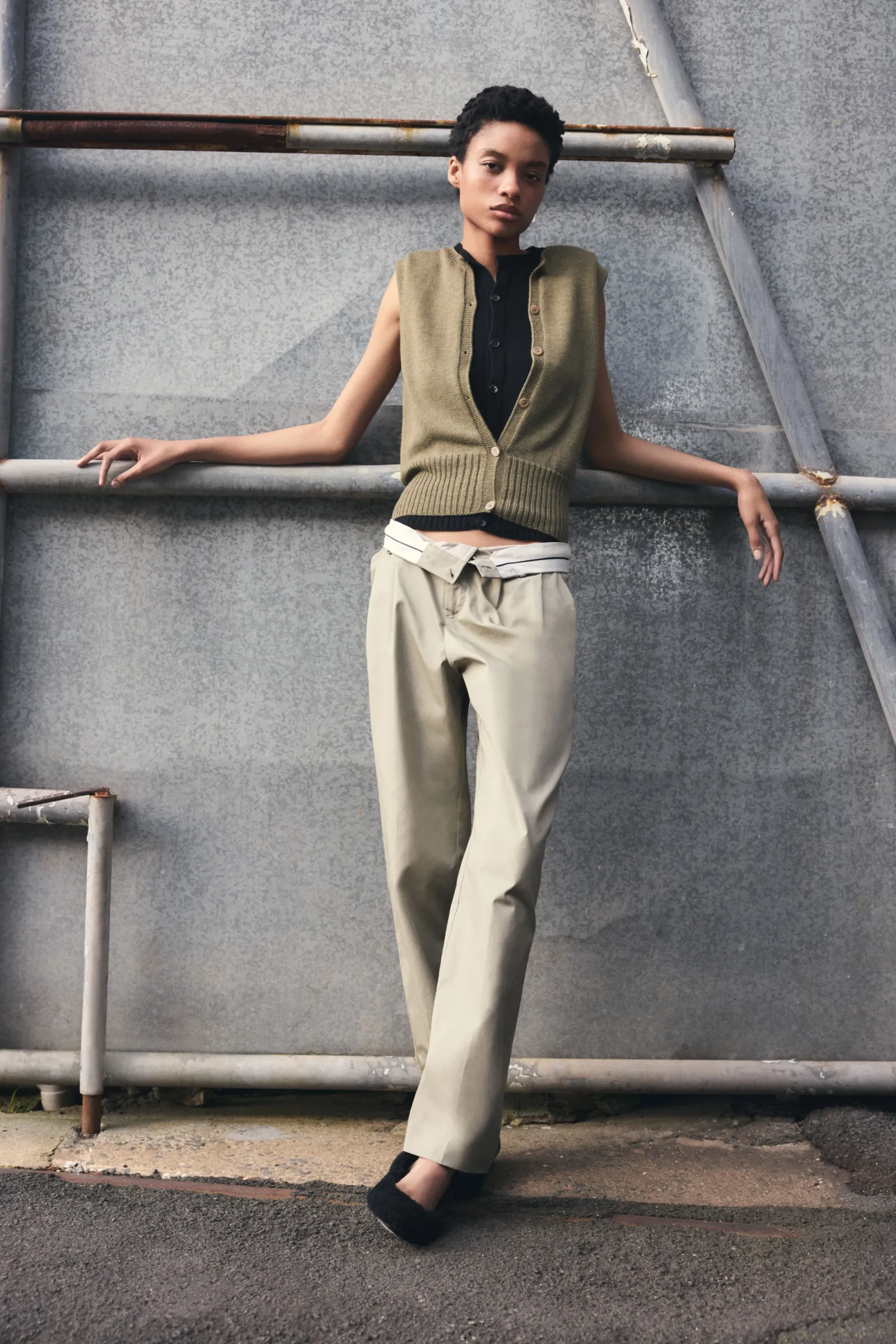 An adult female model poses in an outdoor shot against a concrete wall, wearing an olive vest, black shirt, khaki pants, and black shoes.